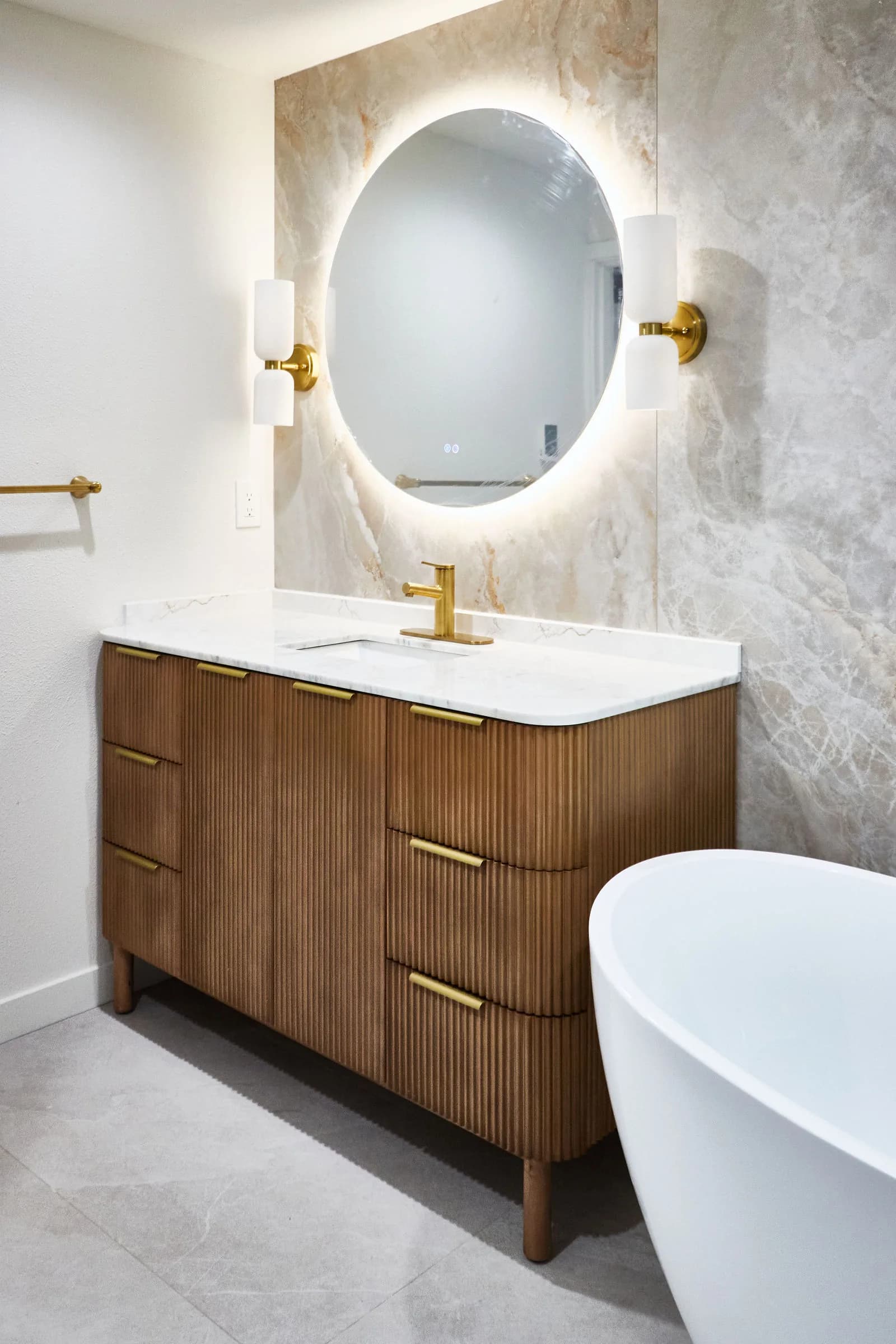 An oval backlit mirror above a walnut vanity, with brass fixtures and a freestanding tub alongside. Bellevue, WA