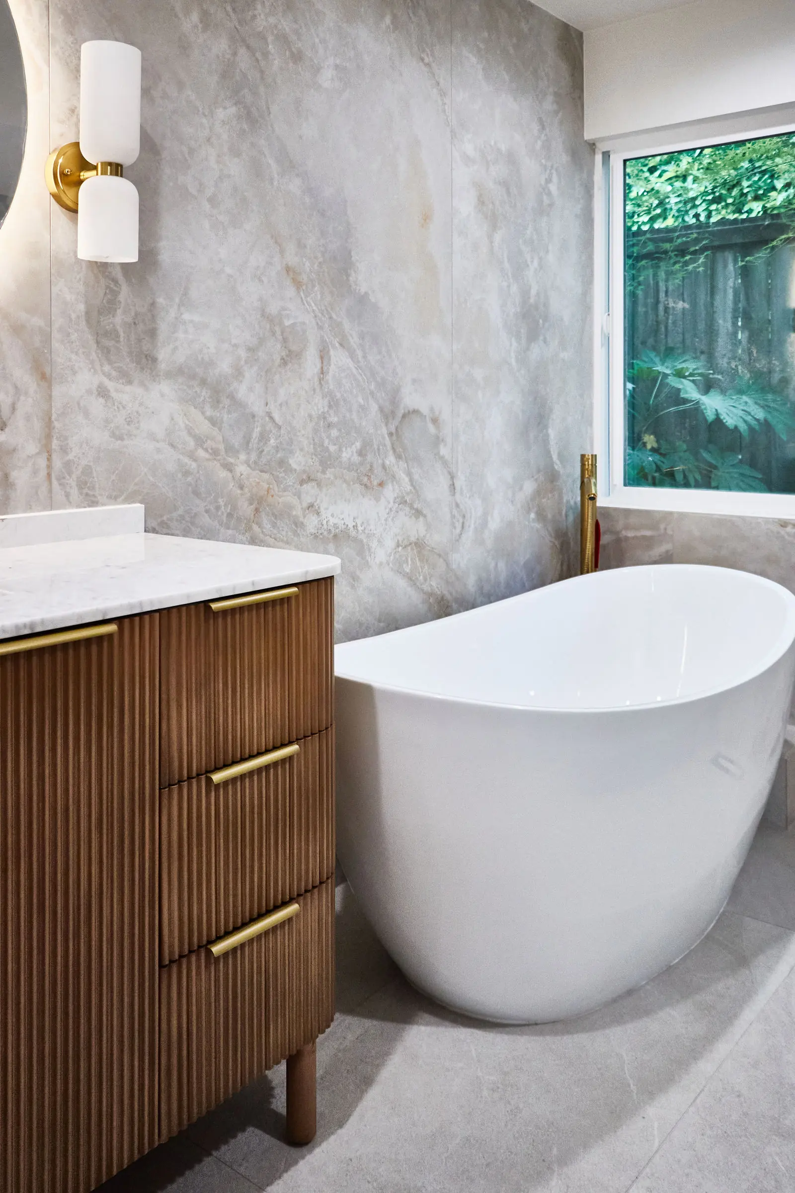 A white freestanding tub and walnut vanity against large-format stone tile, with a window above the tub. Bellevue, WA