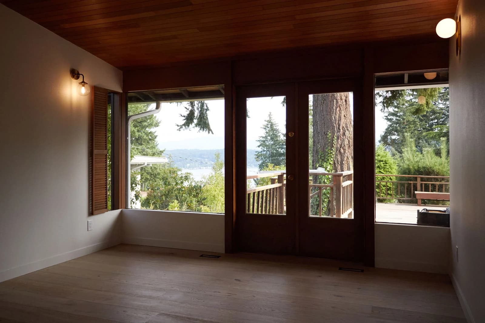 Wood flooring and a wood ceiling frame broad windows and a glass door looking toward the deck and lake. Bellevue, WA