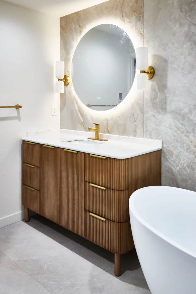 An oval backlit mirror above a walnut vanity, with brass fixtures and a freestanding tub alongside. Bellevue, WA