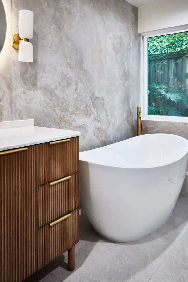 A white freestanding tub and walnut vanity against large-format stone tile, with a window above the tub. Bellevue, WA