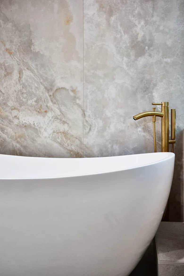 The curved edge of a white freestanding tub beside a brass floor-mounted filler and stone tile. Bellevue, WA